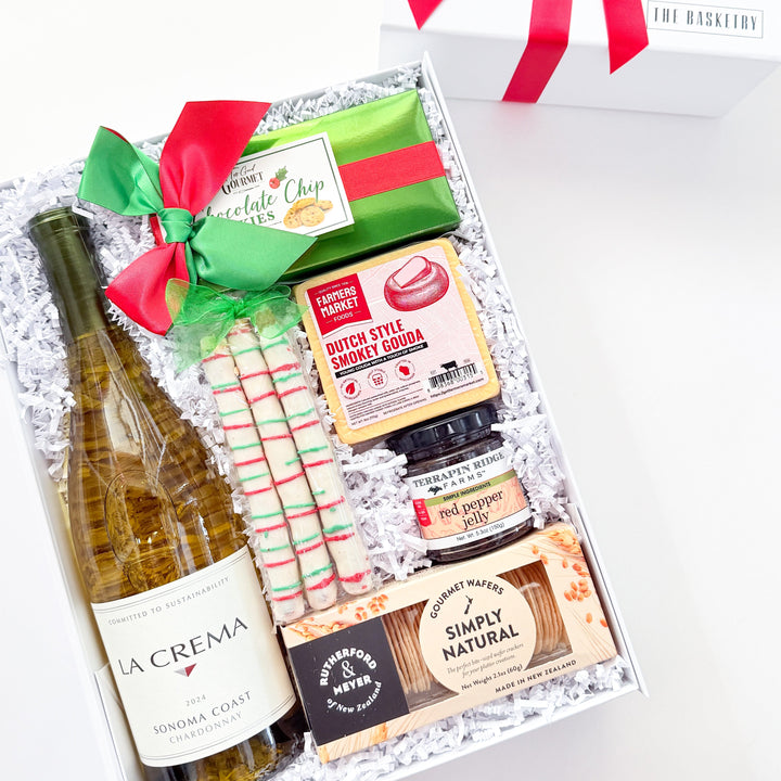 A curated gift box set on a white background, featuring a bottle of La Crema Sauvignon Blanc, a green and red wrapped item, striped candy canes, a jar of preserves, and packaged snacks in white crinkle paper. A white gift box with a red ribbon bow sits behind the open box.