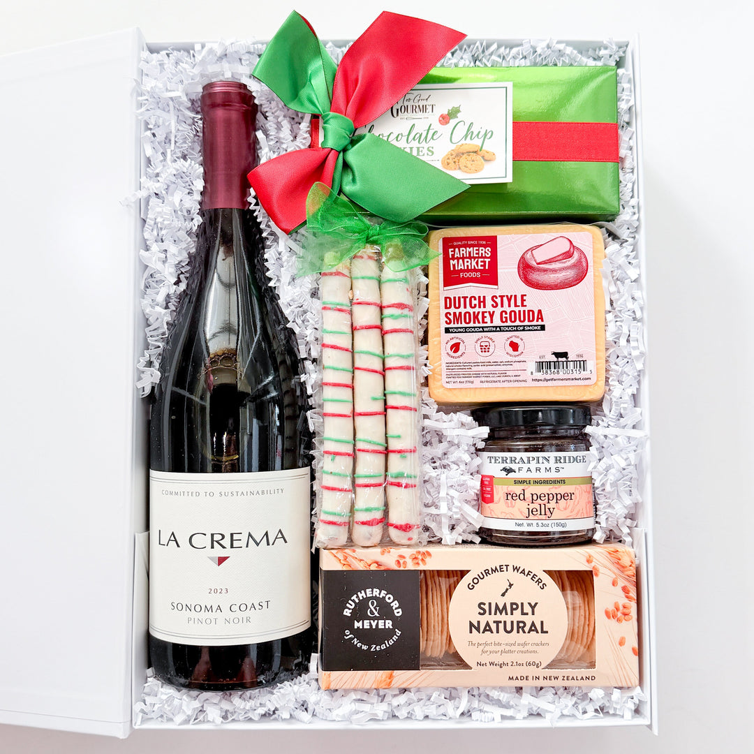 A curated gift box filled with assorted food items, including a bottle of La Crema red wine, a green chocolate bar, Dutch Style Soddy Gouge, red pepper, and Simply Natural crackers, tied with a red and green bow on a white background.