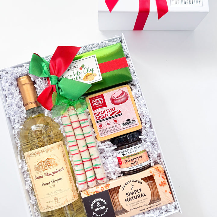 A curated gift box set on a white background, featuring a bottle of wine, candy canes, gourmet snacks, and a white gift box with a red ribbon. The wine bottle label reads &