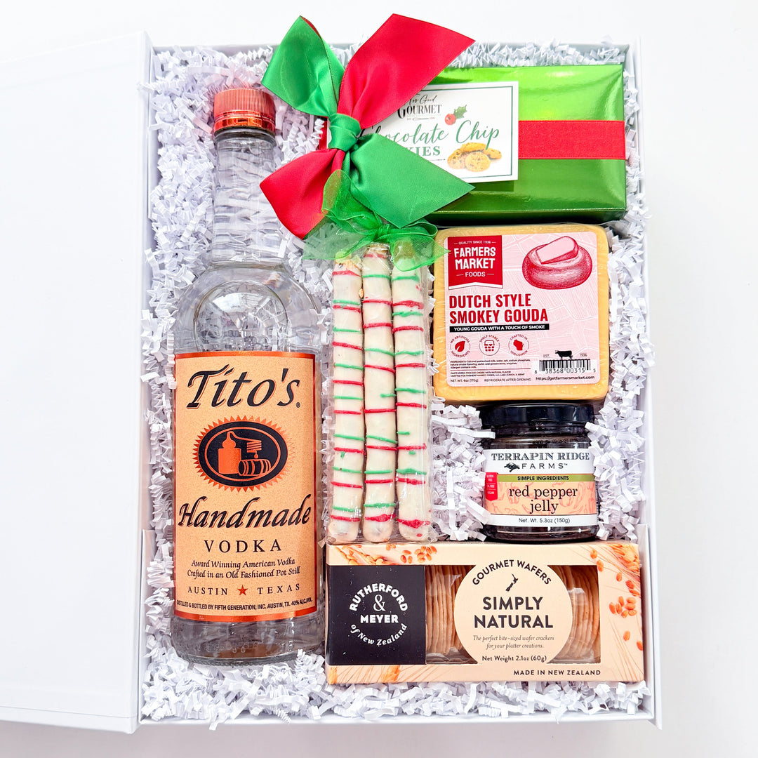 A curated gift box filled with assorted snacks and drinks, tied with a red and green bow. Visible items include a bottle of Tito's Handmade Vodka, a bag of Dutch Style Snackerels, a pack of red and white striped candy canes, a bag of Simply Natural chocolate, and a green bag of coffee chips.