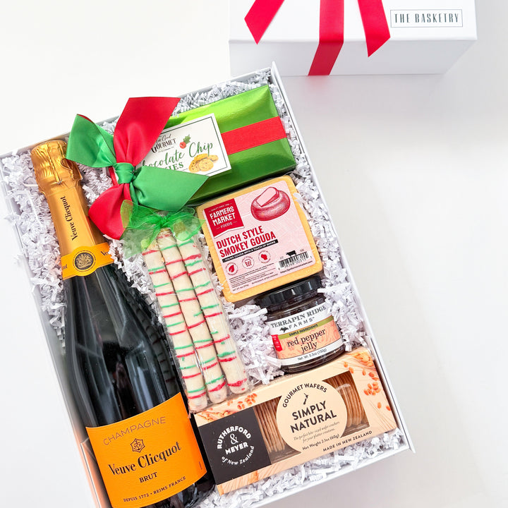 A curated gift box set on a white background, featuring an open box with a bottle of Bréme Clérical champagne, assorted chocolates, candy canes, and a red/green ribbon, alongside a closed white gift box with a red bow. The brand &