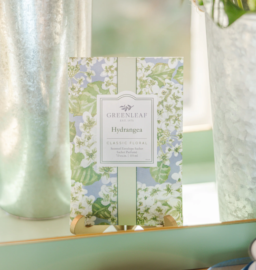 Photograph of a Greenleaf Hydrangea scented envelope sachet positioned on a wooden stand between two galvanized metal buckets, highlighting a watercolor floral pattern of white petals and green leaves against a soft blue background