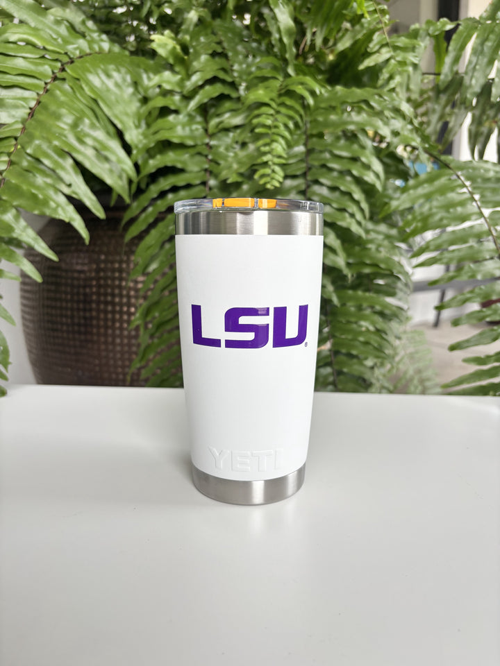 Digital photograph of a white YETI tumbler featuring a purple LSU logo and an orange magnetic slider lid sitting on a white table against a backdrop of a lush green fern and a textured brown pot