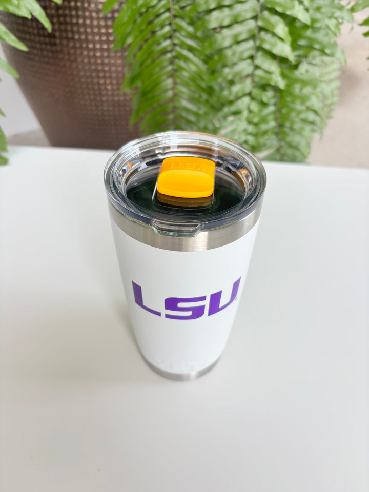 Digital photograph of a white Yeti tumbler featuring a purple LSU logo and a vibrant yellow magnetic slider positioned on a white table against a green fern background