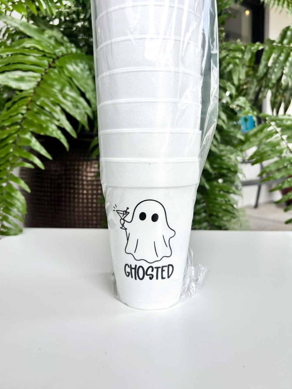 Photograph of a stack of white styrofoam cups wrapped in plastic on a white table featuring a black graphic of a ghost holding a martini glass with the word GHOSTED in front of lush green fern leaves