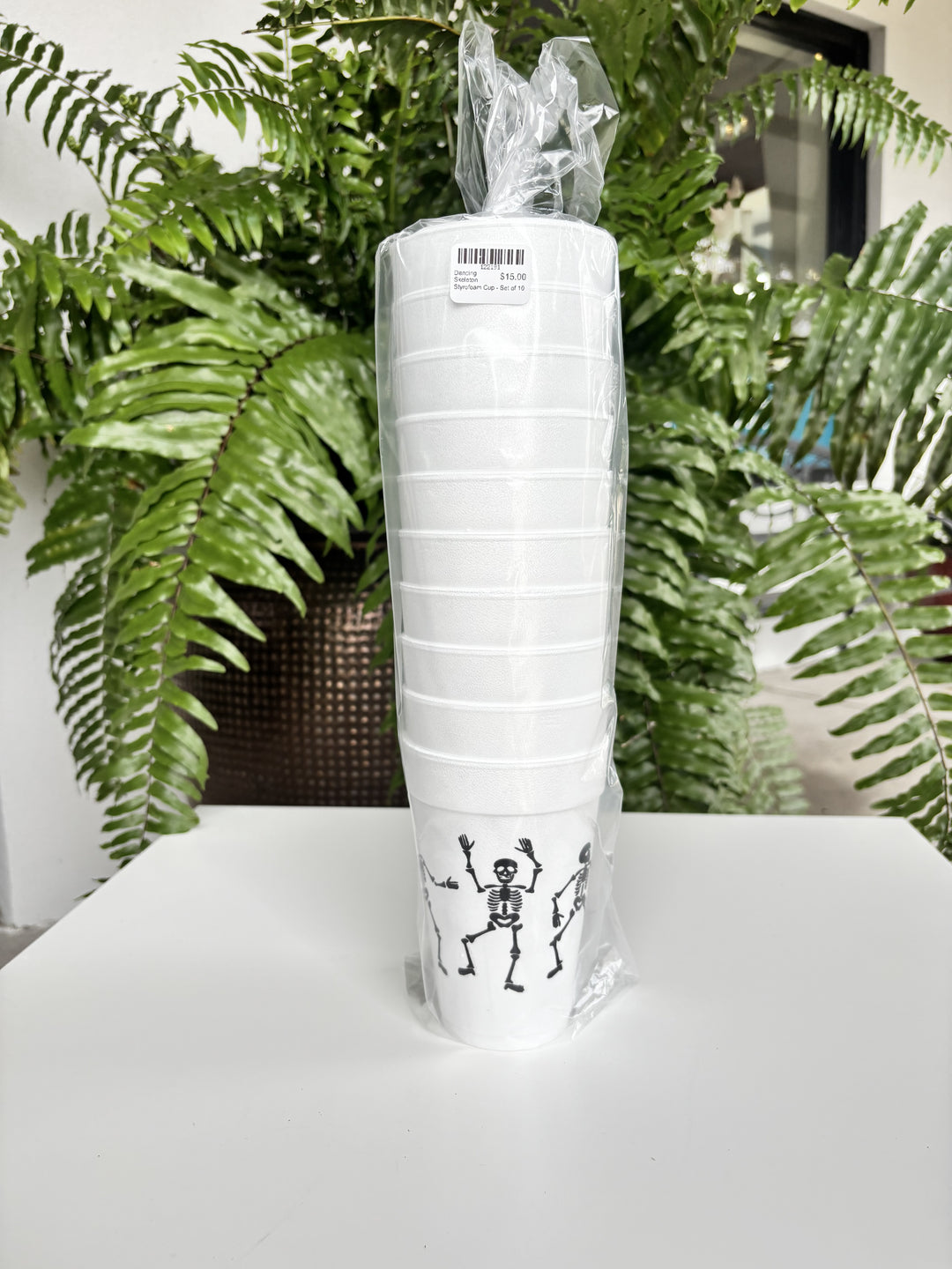 Photograph of a set of ten white styrofoam cups featuring black dancing skeleton illustrations and a barcode label, wrapped in clear plastic and resting on a white table before a lush green fern in a bronze pot