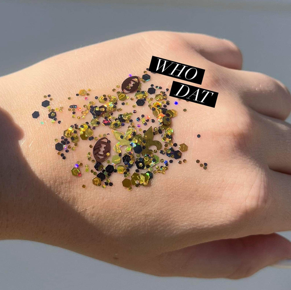 Cosmetic body art of New Orleans Saints themed glitter on a hand featuring a golden fleur-de-lis, brown football sequins, and black hexagonal flakes with WHO DAT text overlay