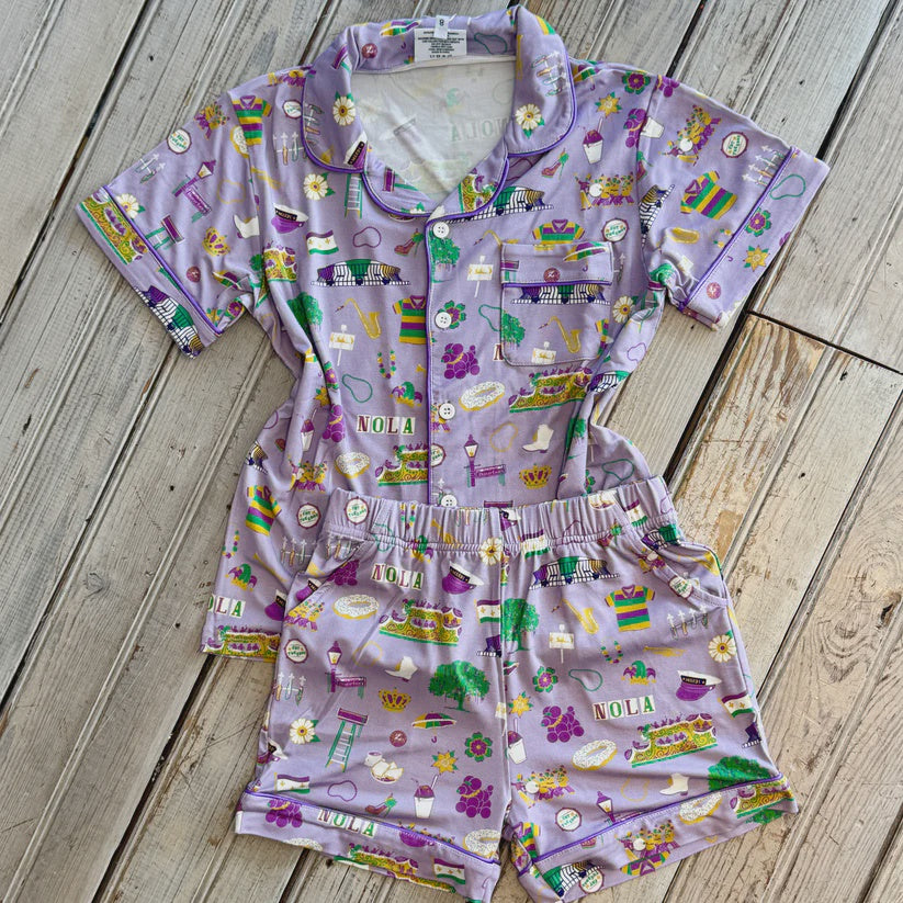 Photograph of a purple New Orleans themed two-piece pajama set laid on a wood floor featuring the Caesars Superdome and streetcars with vibrant illustrations of Mardi Gras parade floats, golden saxophones, and colorful snowballs