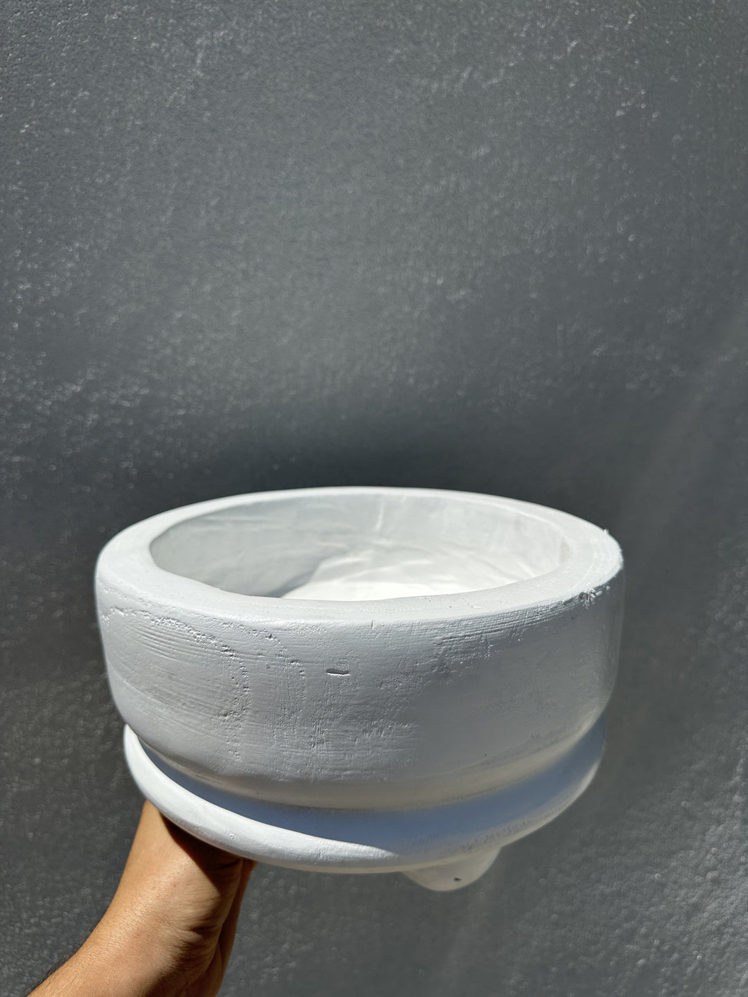 Hand-painted wooden vessel by an unknown artist featuring a white bowl held against a grey textured wall with visible wood grain, a thick rounded rim, and a pedestal base under bright direct sunlight