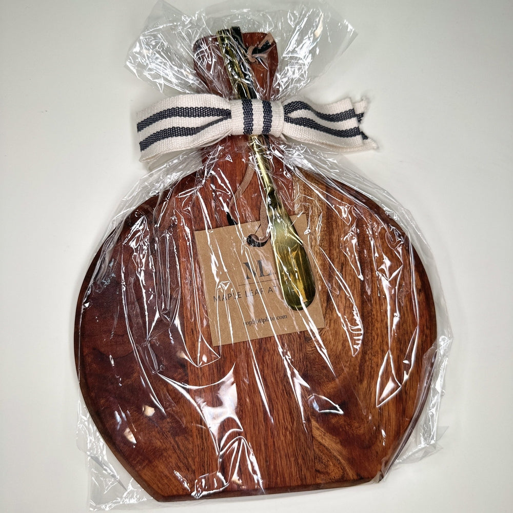 Handcrafted wooden cheese board by Maple Leaf At Home featuring a gold-toned spreader knife and a black and white striped ribbon bow wrapped in clear gift cellophane on a white surface