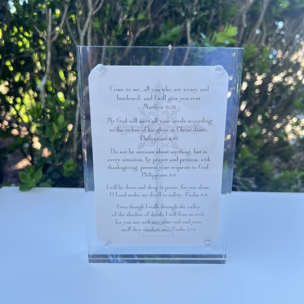 Acrylic block frame plaque featuring Christian Bible verses on a white surface outdoors with a faint ornate cross illustration, elegant serif typography, and a bokeh background of lush sun-dappled green foliage