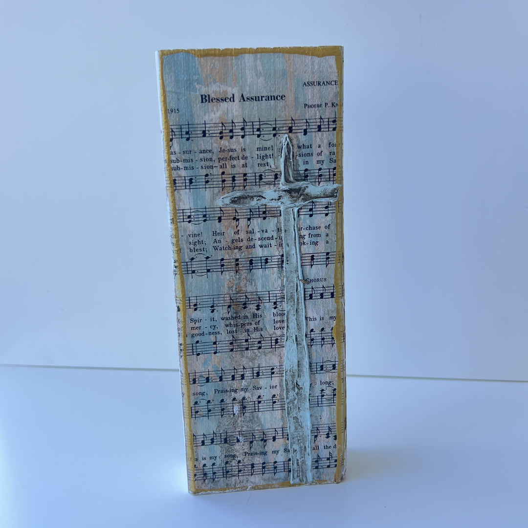 Mixed media decoupage wood block featuring the vintage sheet music for the hymn Blessed Assurance by Phoebe P. Knapp with a raised textured light-blue cross and distressed gold-painted edges centered on a white background