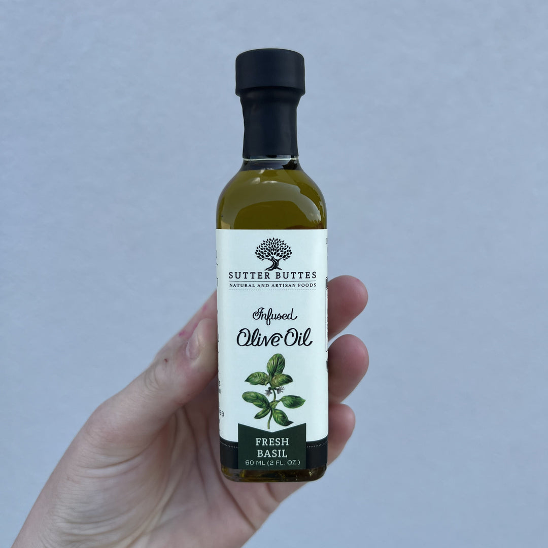 Photograph of Sutter Buttes Natural and Artisan Foods Fresh Basil Infused Olive Oil held in a hand featuring a clear 60 ml bottle with golden liquid, a black cap, and a white label with a tree logo and green leaf illustrations