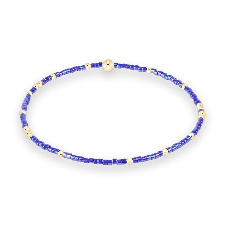 Beaded jewelry stretch bracelet centered on a white background featuring iridescent violet-blue cylindrical beads accented by polished gold-tone spacers and a large central gold sphere