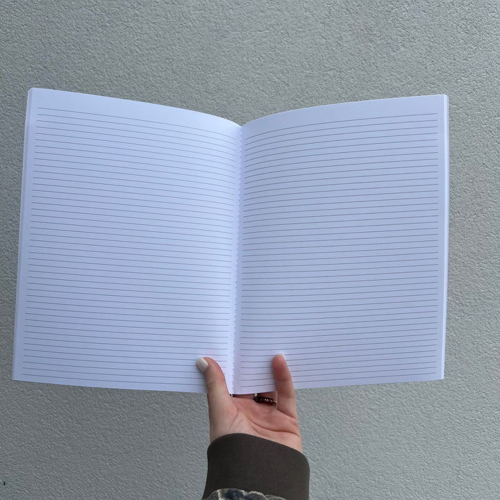 Photograph of an open white-lined notebook held by a hand against a grey textured wall, featuring thin blue horizontal lines, a thumb with pale nail polish, and a dark brown ribbed sleeve