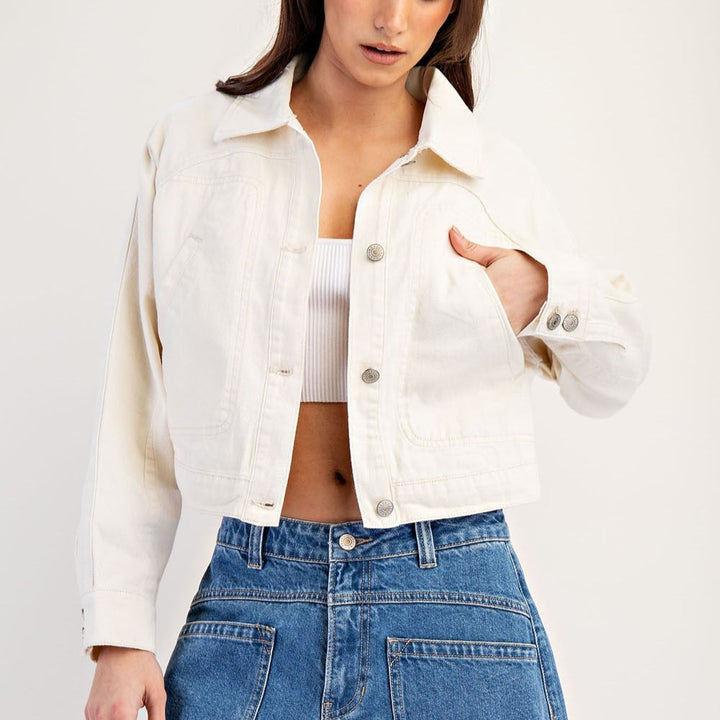 Photograph of a model showcasing a cropped white denim jacket and blue utility jeans featuring large front pockets and metallic buttons in a studio setting