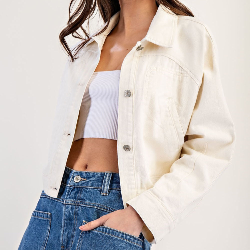 Fashion photograph of a woman modeling an off-white denim jacket featuring silver-toned buttons and panel stitching paired with a white ribbed crop top and blue jeans in a studio setting