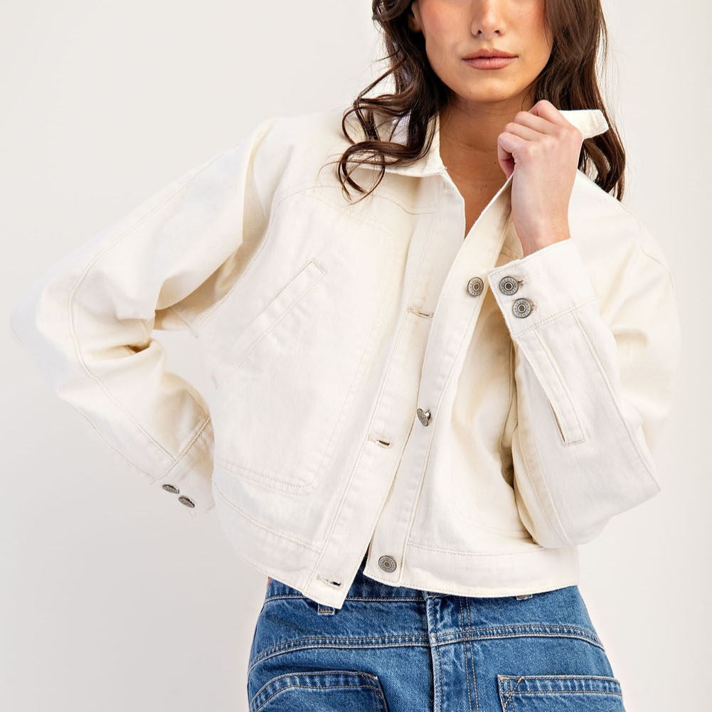 Studio fashion photograph of a woman wearing an off-white cropped denim jacket featuring metallic buttons and visible panel stitching paired with blue jeans in a neutral studio setting