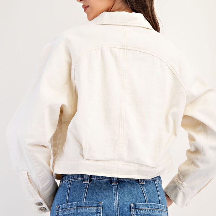 Studio photograph of a woman from behind in a minimalist studio wearing an off-white cropped denim jacket and blue jeans featuring prominent vertical stitching and buttoned cuffs