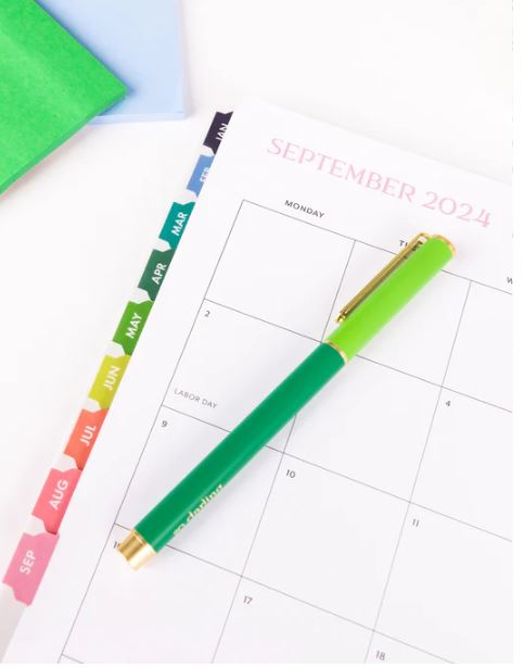 Photograph of a Darling brand stationery set featuring a September 2024 calendar page on a white desk with a two-tone green pen, gold metallic accents, and a vibrant row of multi-colored monthly index tabs