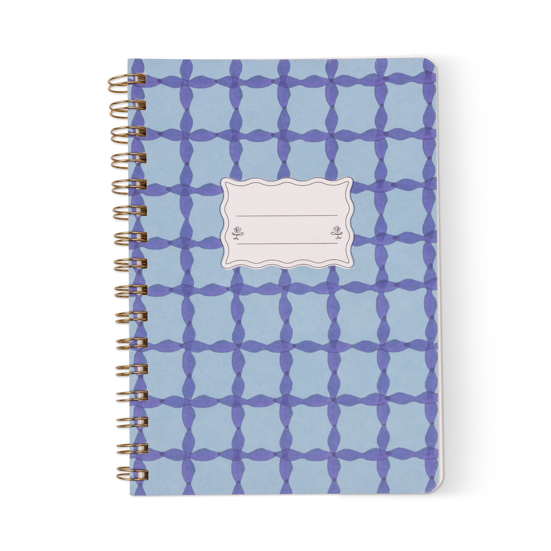 Stationery spiral-bound notebook featuring a blue and purple cross-patterned cover with gold-tone wire binding and a scalloped white nameplate centered on a white background