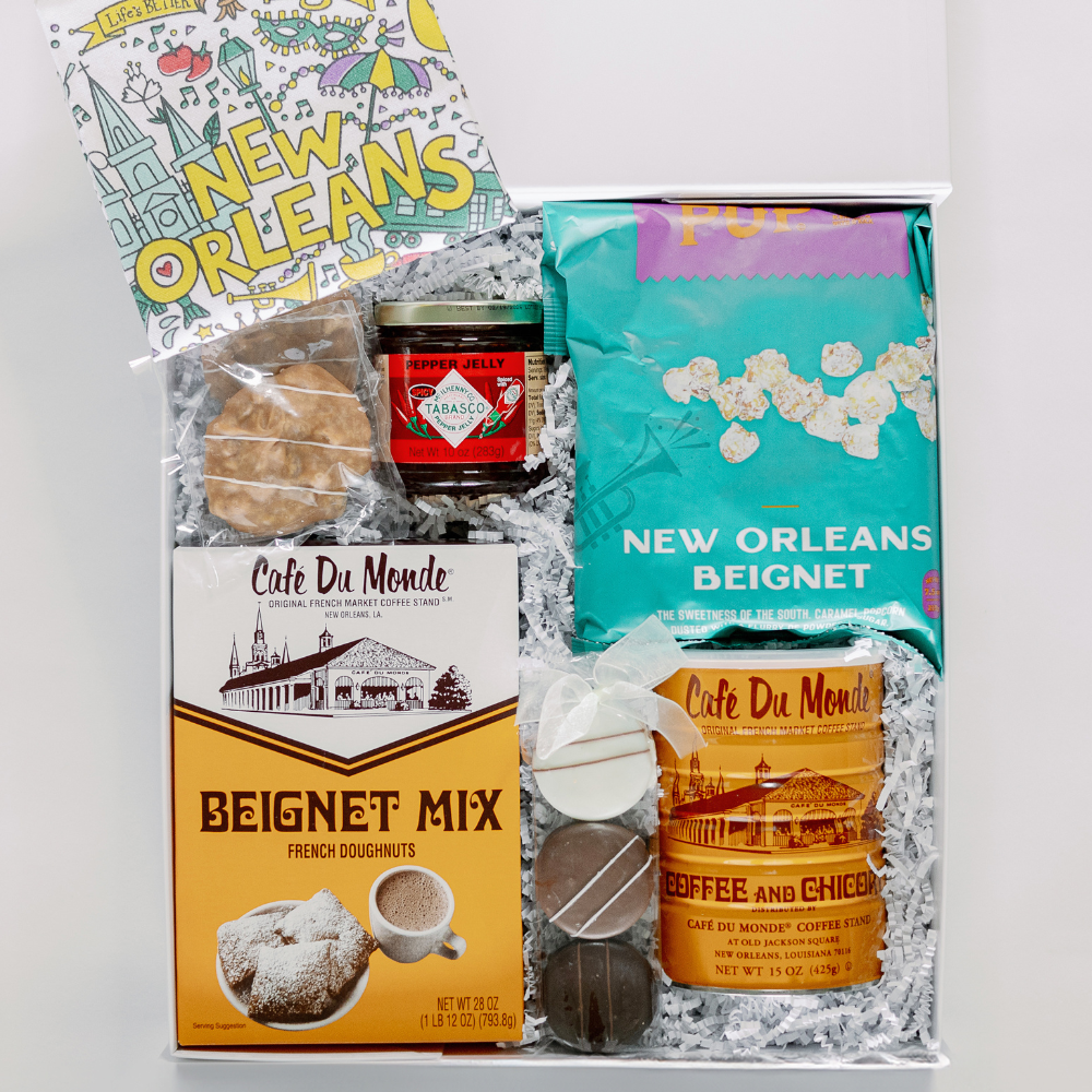 Photograph of a curated New Orleans gift box featuring a yellow box of Cafe Du Monde beignet mix, a vibrant teal bag of beignet popcorn, and a jar of red Tabasco pepper jelly alongside illustrations of the St. Louis Cathedral and Jackson Square