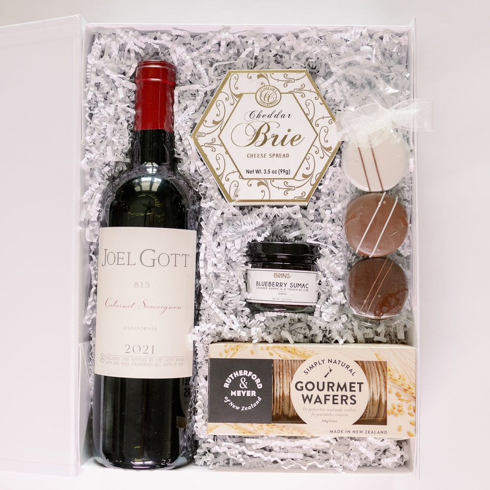 Photograph of a gourmet gift hamper in a white box featuring a bottle of Joel Gott 815 Cabernet Sauvignon, Rutherford & Meyer wafers, and a gold hexagonal box of cheddar brie spread nestled in white shredded paper
