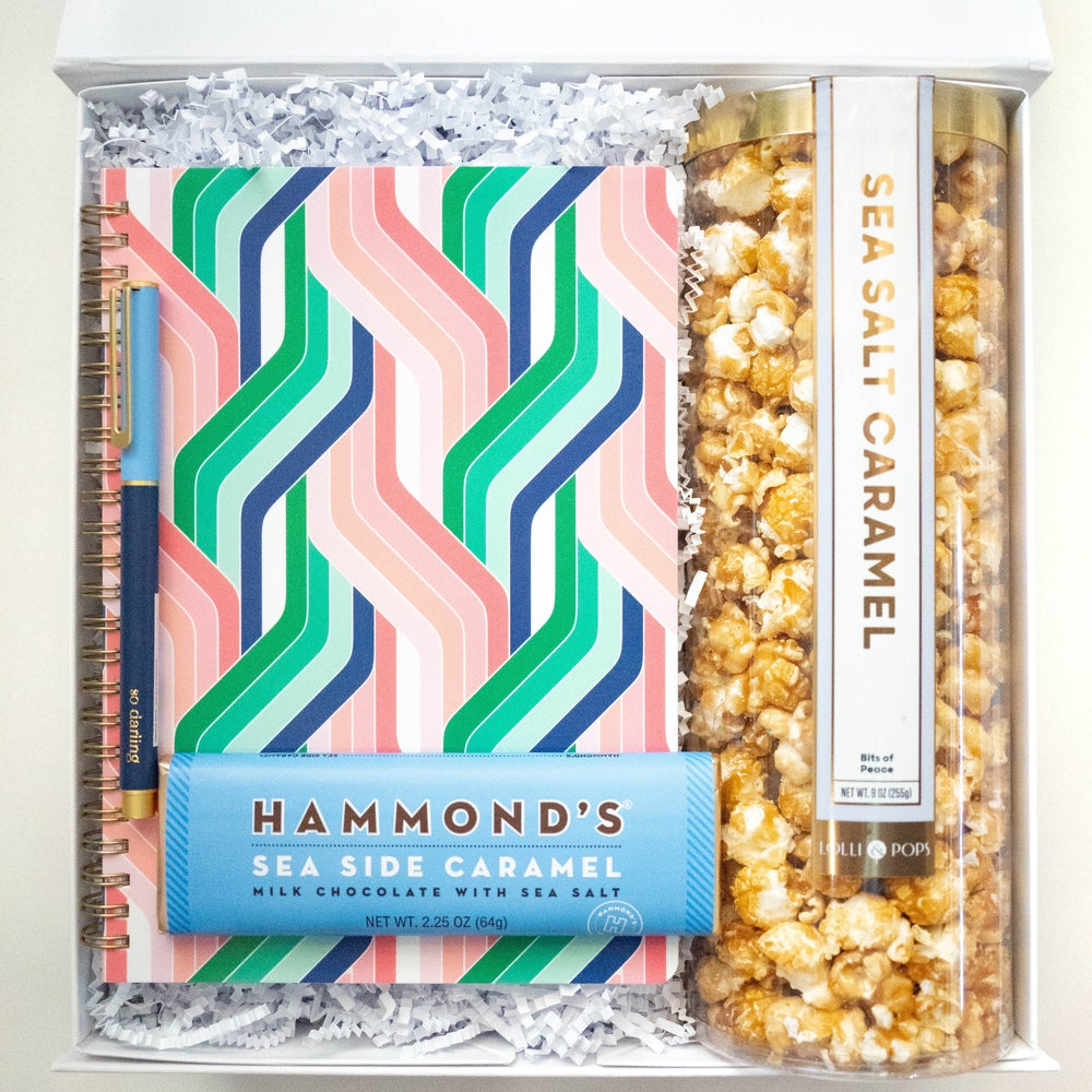 Photograph of a curated gift box featuring a vibrant geometric zigzag notebook, a blue Hammond's Sea Side Caramel chocolate bar, and a tall transparent tube of golden sea salt caramel popcorn nestled in white shredded paper filler
