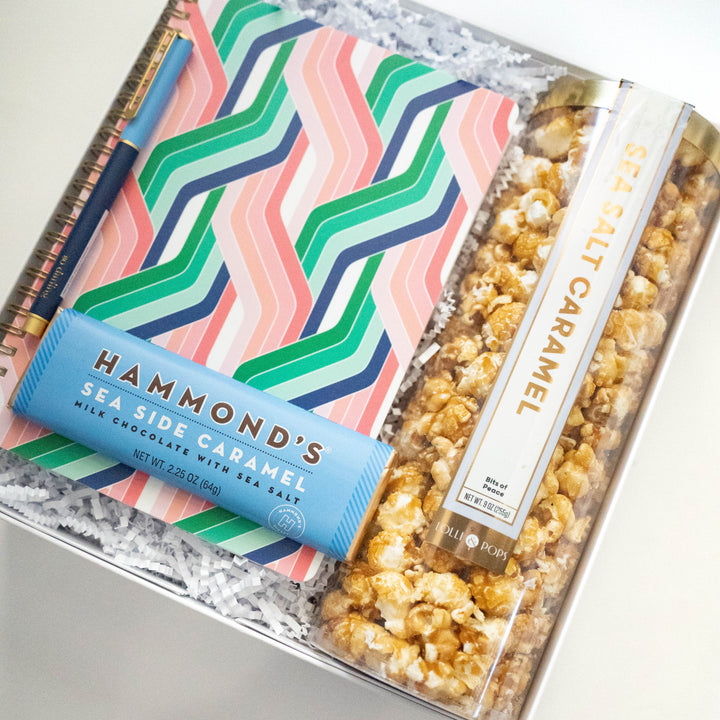 Digital photograph of a curated gift box showcasing a Hammond’s Sea Side Caramel chocolate bar, a tube of gourmet sea salt caramel popcorn, and a vibrant pink and green zigzag patterned notebook paired with a gold-trimmed navy pen nestled in white crinkle paper