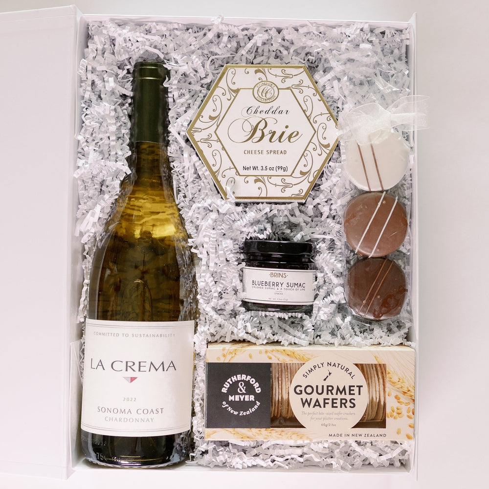Commercial photograph of a luxury wine and gourmet food gift hamper featuring a bottle of La Crema Sonoma Coast Chardonnay, a gold-accented hexagonal box of brie, and three striped chocolate-covered cookies nestled in white shredded paper