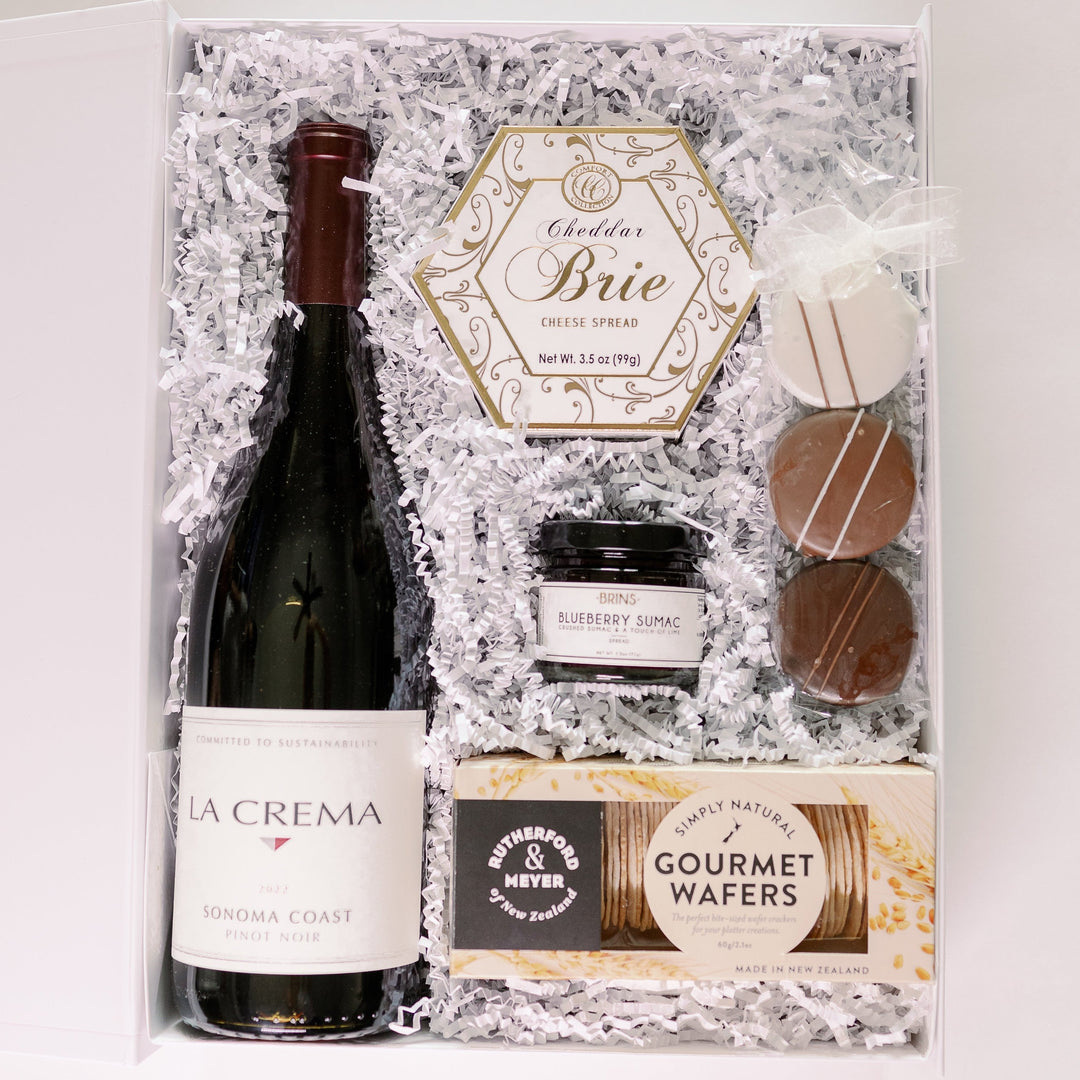 A studio photograph of a gourmet gift box featuring a bottle of La Crema Sonoma Coast Pinot Noir, a hexagonal gold-foiled box of Cheddar Brie, and Rutherford & Meyer wafers nestled in white crinkle paper with three striped chocolate cookies and a jar of blueberry sumac jam