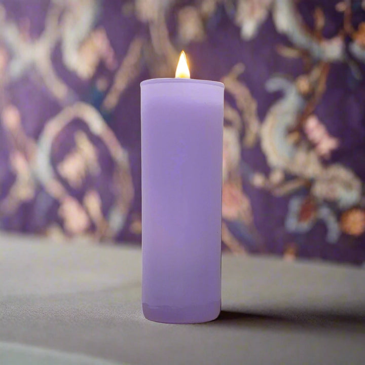 Photograph of a tall lit purple pillar candle on a flat neutral surface featuring a small glowing yellow flame and a blurred violet floral patterned backdrop