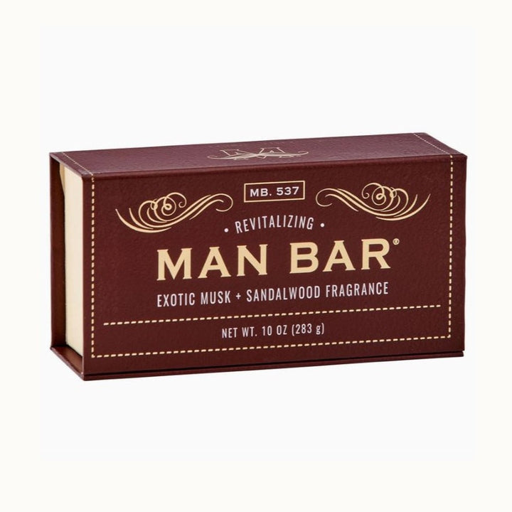 Studio product photograph of a rectangular MAN BAR soap box for Exotic Musk and Sandalwood fragrance on a white background featuring deep burgundy textured packaging, gold-colored ornamental scrolls, and cream-toned dashed border accents