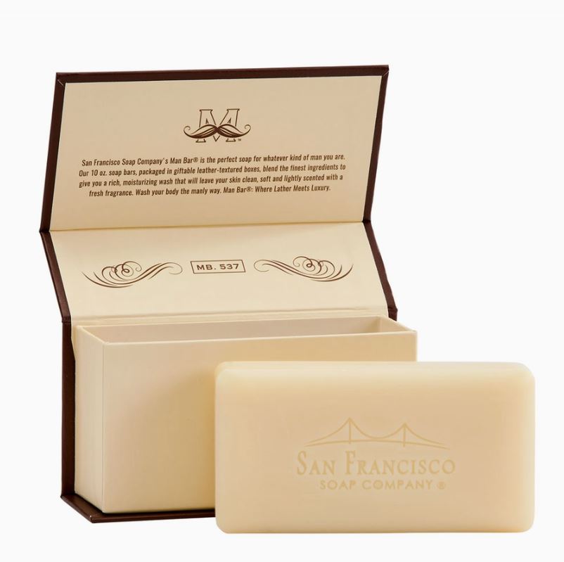 Product photography of the San Francisco Soap Company Man Bar featuring an embossed Golden Gate Bridge on a cream soap bar resting in front of an open brown gift box with a mustache logo and elegant vintage script