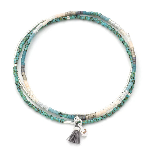Handcrafted glass bead wrap bracelet by Scout featuring three layers of turquoise, cream, and silver seed beads set against a white background with a small grey tassel and a silver logo tag