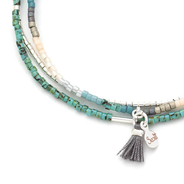 Handcrafted beaded jewelry by Scout Curated Wearables featuring a multi-strand bracelet on a white background with turquoise glass beads, silver metallic accents, and a grey fringe tassel with a branded Scout logo tag