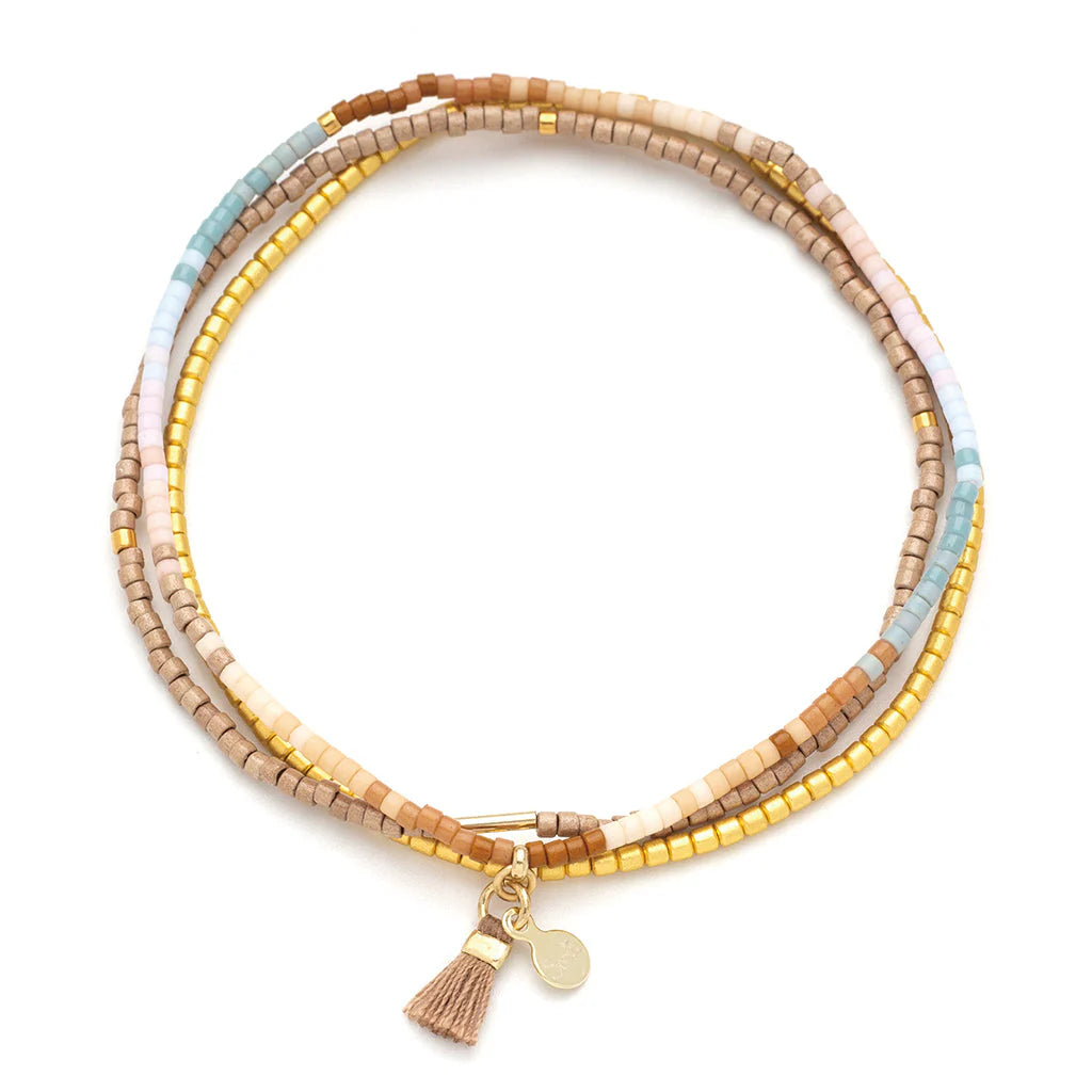 Beaded jewelry by Lotus and Luna featuring a multi-strand wrap bracelet on a white background with muted earth-tone seed beads, metallic gold accents, and a tiny tan tassel charm