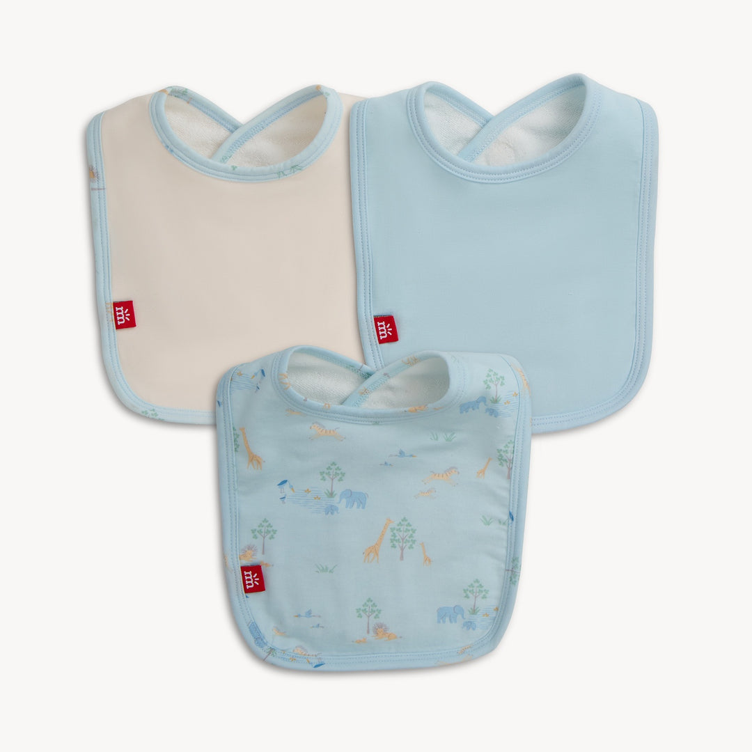 Product photograph of three baby bibs in light blue and cream featuring a safari animal pattern with giraffes and elephants, red fabric logo tags, and soft light blue piping on a white background