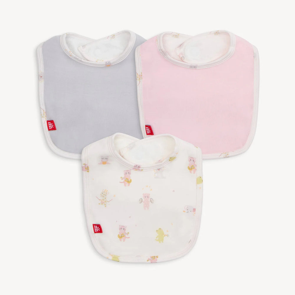 Product photograph of three cotton baby bibs by Mothercare in solid gray, pastel pink, and a white animal musician print featuring signature red brand tags and whimsical illustrations on a clean white background