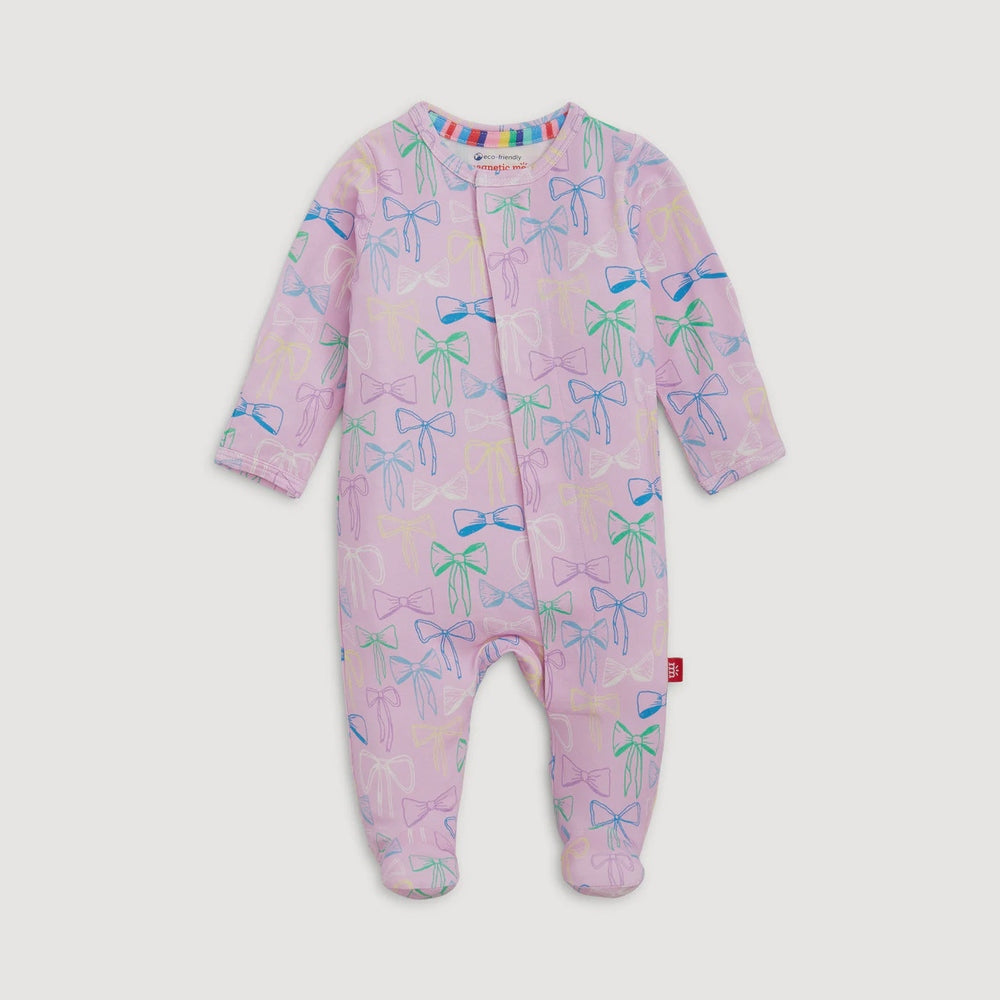Photograph of a Magnetic Me baby footie on a plain grey background featuring a lavender fabric with a pattern of teal, blue, and pink hand-drawn bows and the brand's logo visible on the inner neckline
