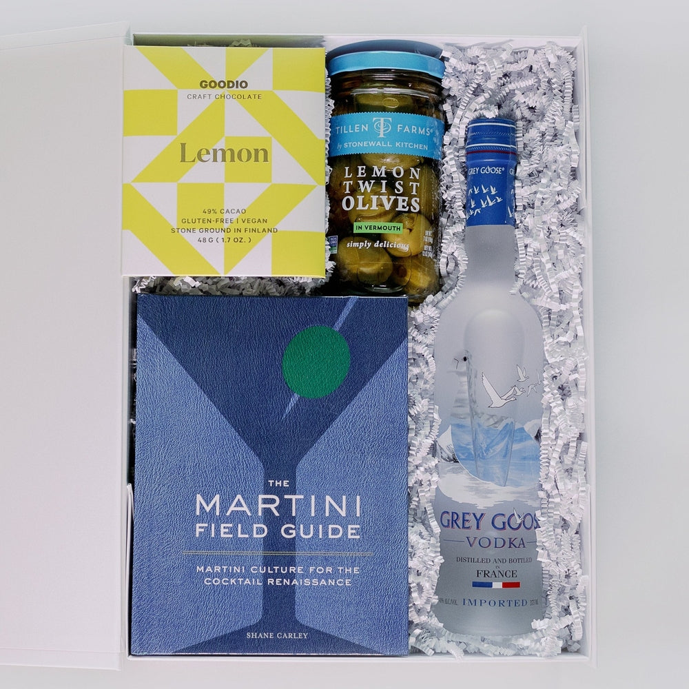 Photograph of a curated martini gift box featuring a book by Shane Carley and Grey Goose vodka from France showcasing a bright yellow geometric wrapper and green lemon twist olives nestled in white shredded paper
