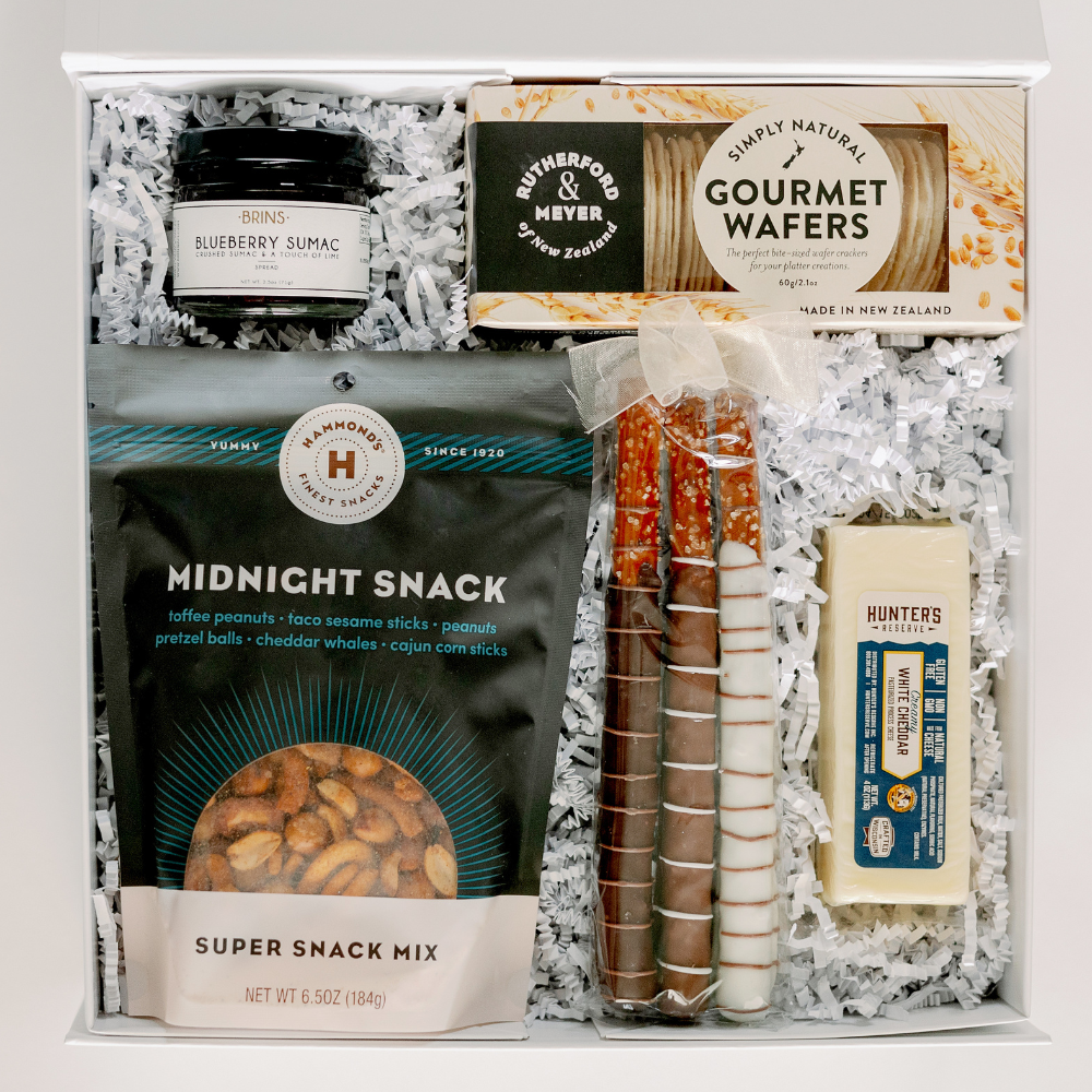 Still life photograph of a curated gourmet gift box featuring Brins Blueberry Sumac spread, Rutherford & Meyer wafers, and Hammond’s Midnight Snack mix in teal packaging arranged in a white container with chocolate-dipped pretzel rods tied with a sheer white bow and Hunter’s Reserve white cheddar cheese on white crinkle paper