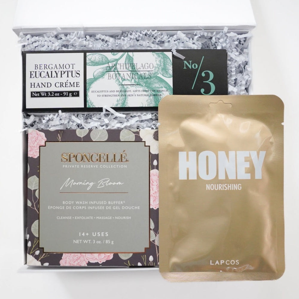 Digital photograph of a curated spa gift box featuring brands Spongellé, LAPCOS, and Archipelago Botanicals alongside a shimmering gold honey mask and floral-patterned packaging nestled in white crinkle paper