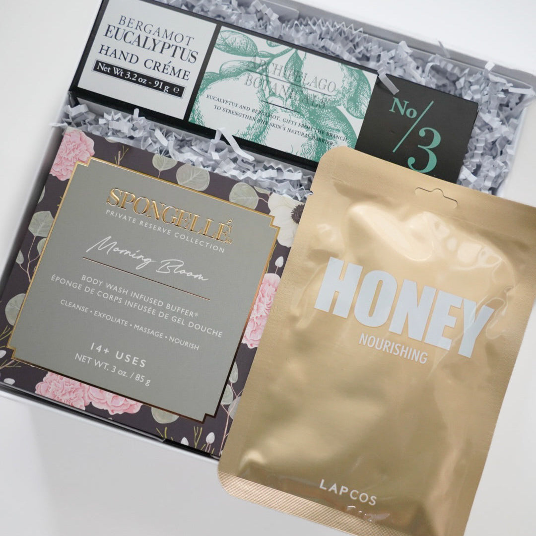 Photograph of a luxury beauty gift box featuring Archipelago Botanicals bergamot eucalyptus hand cream, a floral Spongellé Morning Bloom buffer, and a metallic gold LAPCOS honey mask resting on white crinkle paper