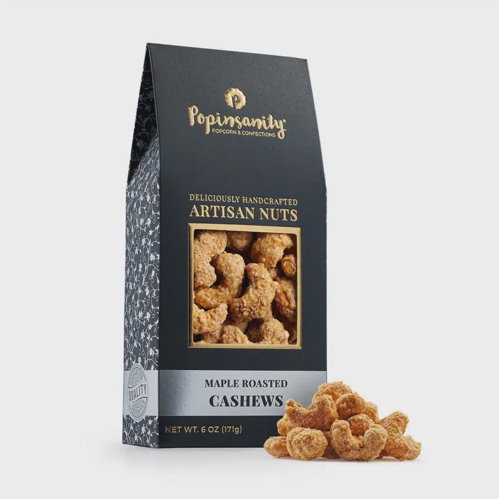 Product photograph by Popinsanity of maple roasted artisan cashews in a premium black box on a white background featuring the gold foil logo, a cluster of crunchy glazed nuts, and a white floral vine pattern on the side panel