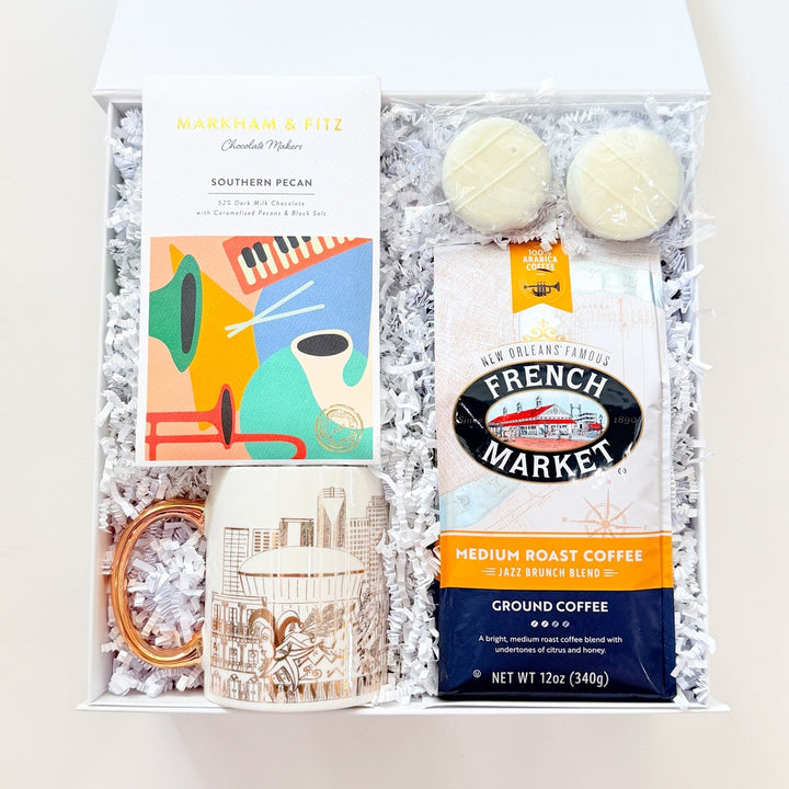 A curated gift box set containing a Mariah &amp; Fitt Southern Pecan box, a bag of French Market ground coffee, two cream macarons, a ceramic mug, and a small gold ring, all arranged in white crinkle paper inside an open white gift box.