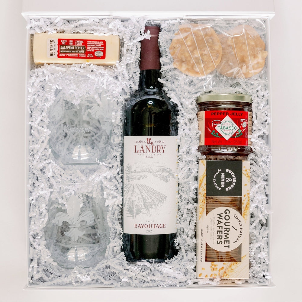 Commercial product photograph of a Landry Vineyards gourmet gift set featuring a bottle of Bayoutage wine, two glasses etched with the iconic Fleur-de-lis symbol, and a jar of Tabasco pepper jelly nestled in white shredded paper filler inside a white box