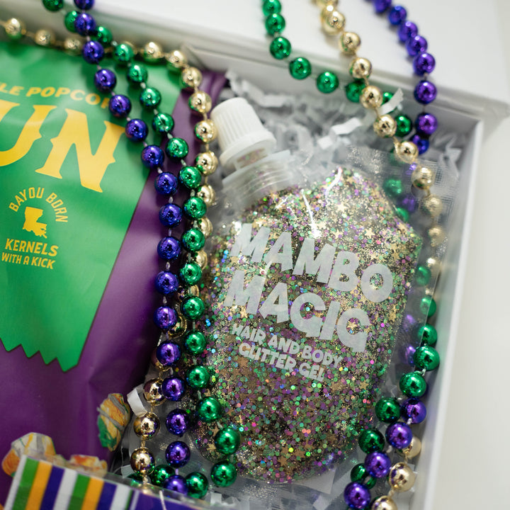 Photograph of a Mardi Gras themed gift box featuring a pouch of Mambo Magic hair and body glitter gel, shimmering purple, green, and gold beads, and a Bayou Born popcorn bag displaying the Louisiana state map icon