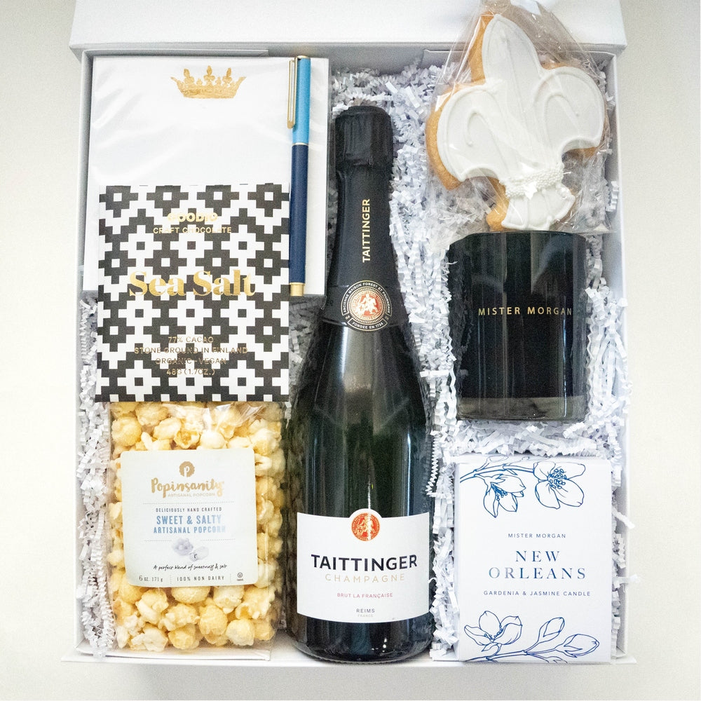 Photograph of a New Orleans themed gift box featuring a bottle of Taittinger Champagne, a white-iced fleur-de-lis cookie, and a gold crown card nestled in white shredded paper