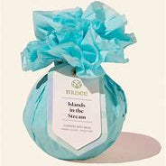 Product photograph of a Musee bath bomb titled Islands in the Stream wrapped in ruffled turquoise tissue paper with a pointed white tag on a neutral cream background